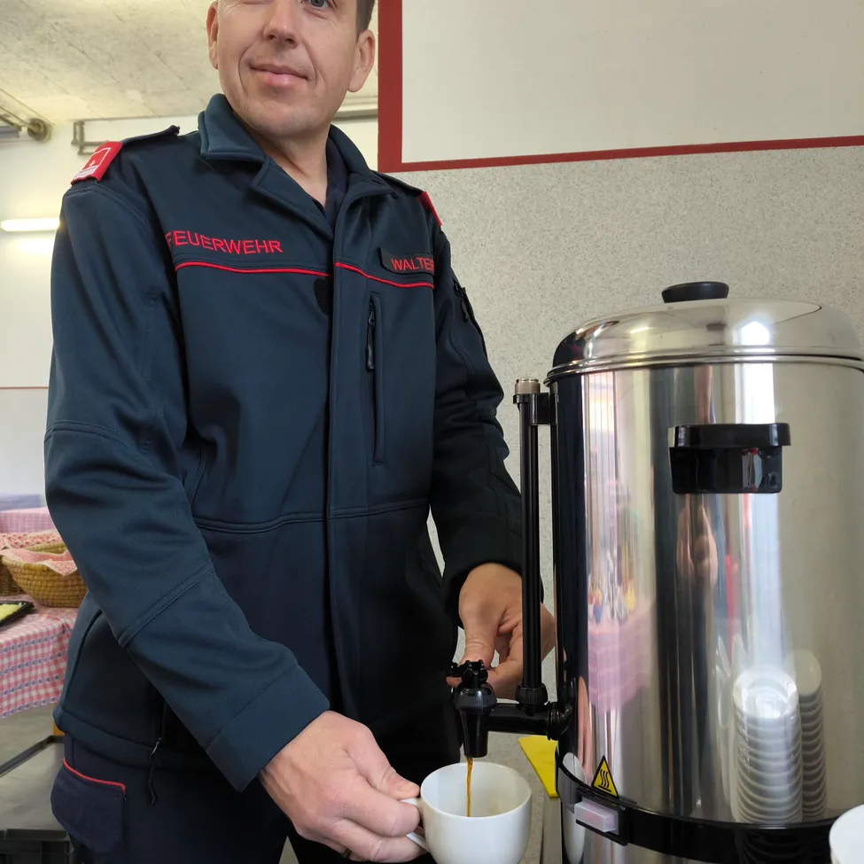 Ein Feuerwehrmann füllt einen Becher mit Kaffee aus einem großen silbernen Spender. Er trägt einen dunkelblauen Uniformmantel mit rotem Saum und dem Wort 'Feuerwehr' auf der linken Seite.