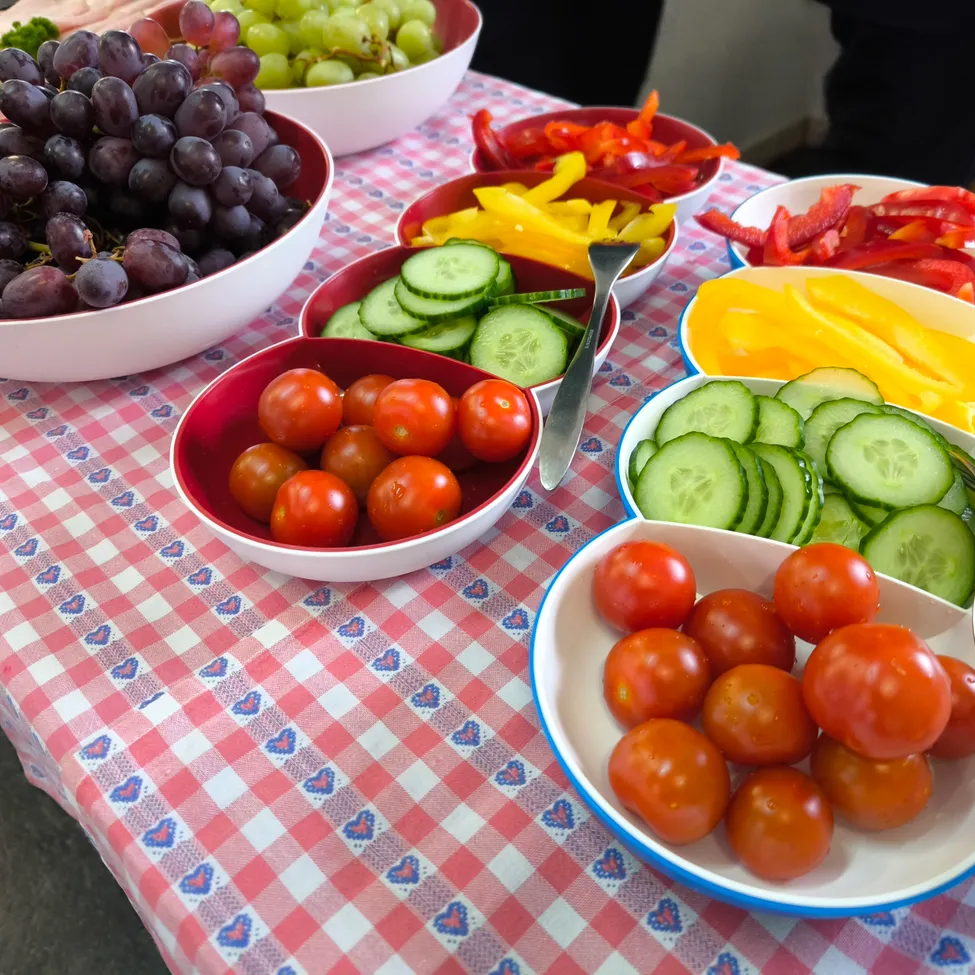 Ein Tisch mit rot-weiß kariertem Tischtuch ist mit verschiedenen Obst- und Gemüsesorten wie Trauben, Tomaten und Gurken beladen. Schüsseln mit roten und gelben Paprikaschoten sind ebenfalls vorhanden.