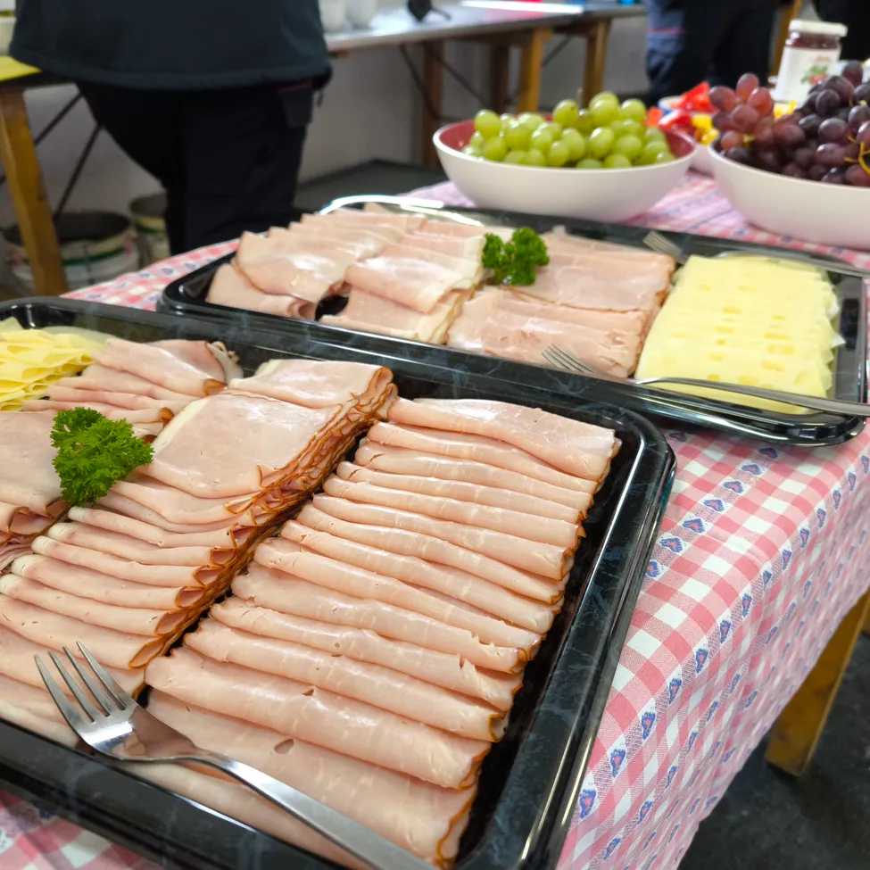 Ein Tisch mit einer pink-weißen Tischdecke hat zwei Tabletts mit geschnittenem Schinken, Käse und einer Schüssel Trauben. Eine Gabel und ein Messer liegen auf den Tabletts. Im Hintergrund steht eine Person in der Nähe eines Tisches mit einer Schüssel Obst und einem Glas.