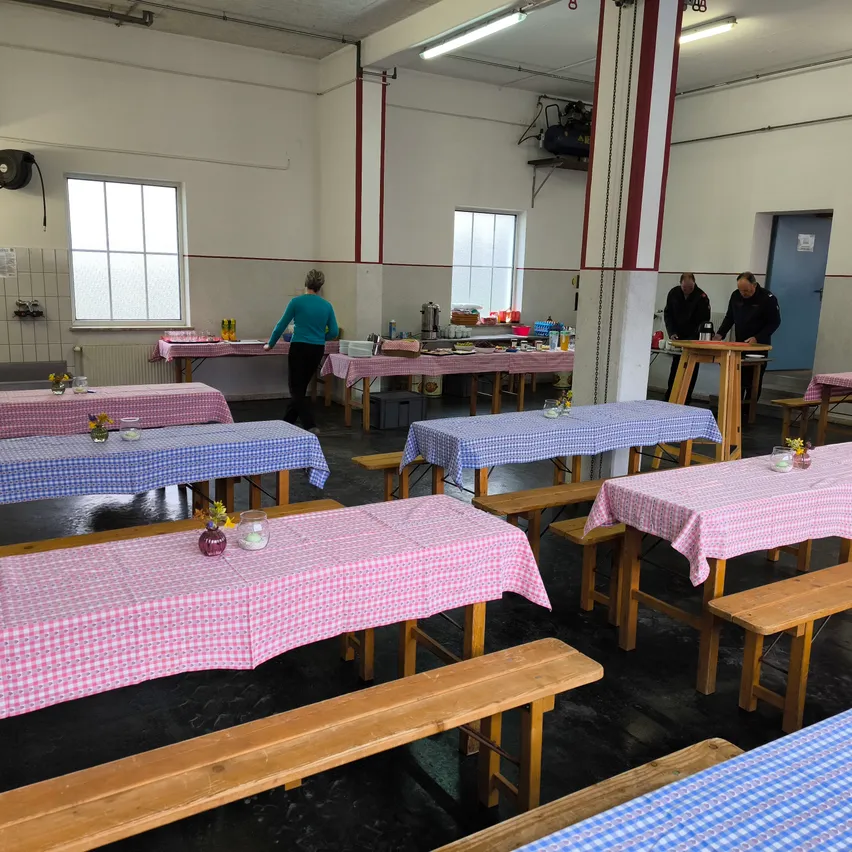 Ein Raum mit mehreren Holztischen und Bänken. Alle Tische sind mit pink-blau karierten Tischdecken bedeckt. Zwei Personen stehen in der Nähe eines Holztisches, möglicherweise beim Essen vorbereiten.