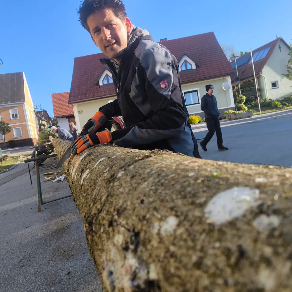 Ein Mann sägt einen Baumstamm mit einer Kettensäge auf der Straße. Im Hintergrund sind Häuser mit Solarpanelen und eine Person, die geht.