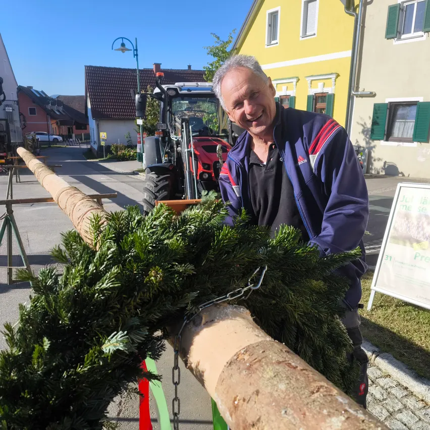 Ein Mann steht mit einem großen Tannenbaum, lächelt vor einem gelben Haus, mit einem Traktor in der Nähe.