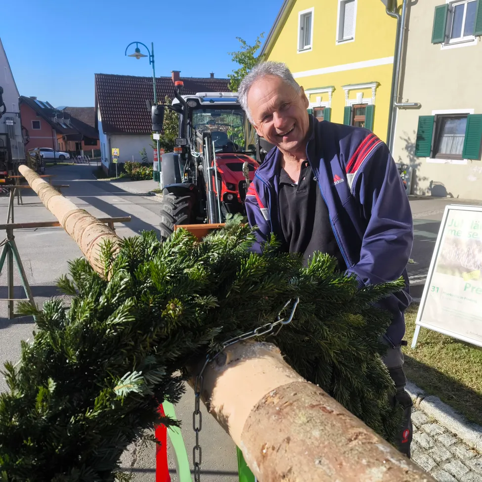 Ein Mann steht mit einem großen Tannenbaum, lächelt vor einem gelben Haus, mit einem Traktor in der Nähe.
