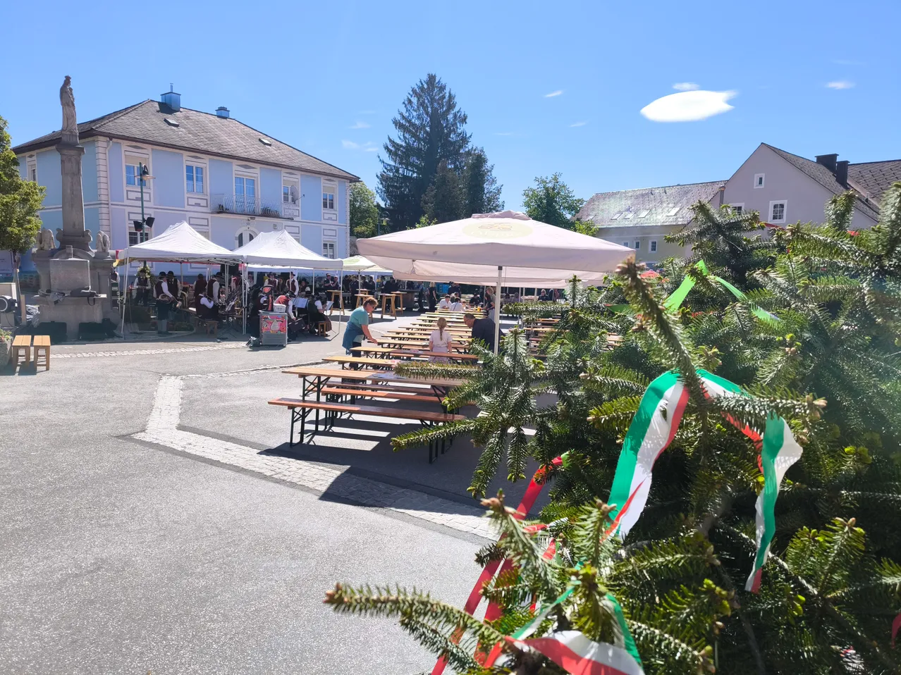 Ein Freiluftbereich mit einer großen Gruppe von Menschen, Bänken, Tischen und weißen Sonnenschirmen. Ein hoher Baum mit grünen, roten und weißen Bändern ist im Vordergrund. Ein weißes Gebäude und Bäume sind im Hintergrund.