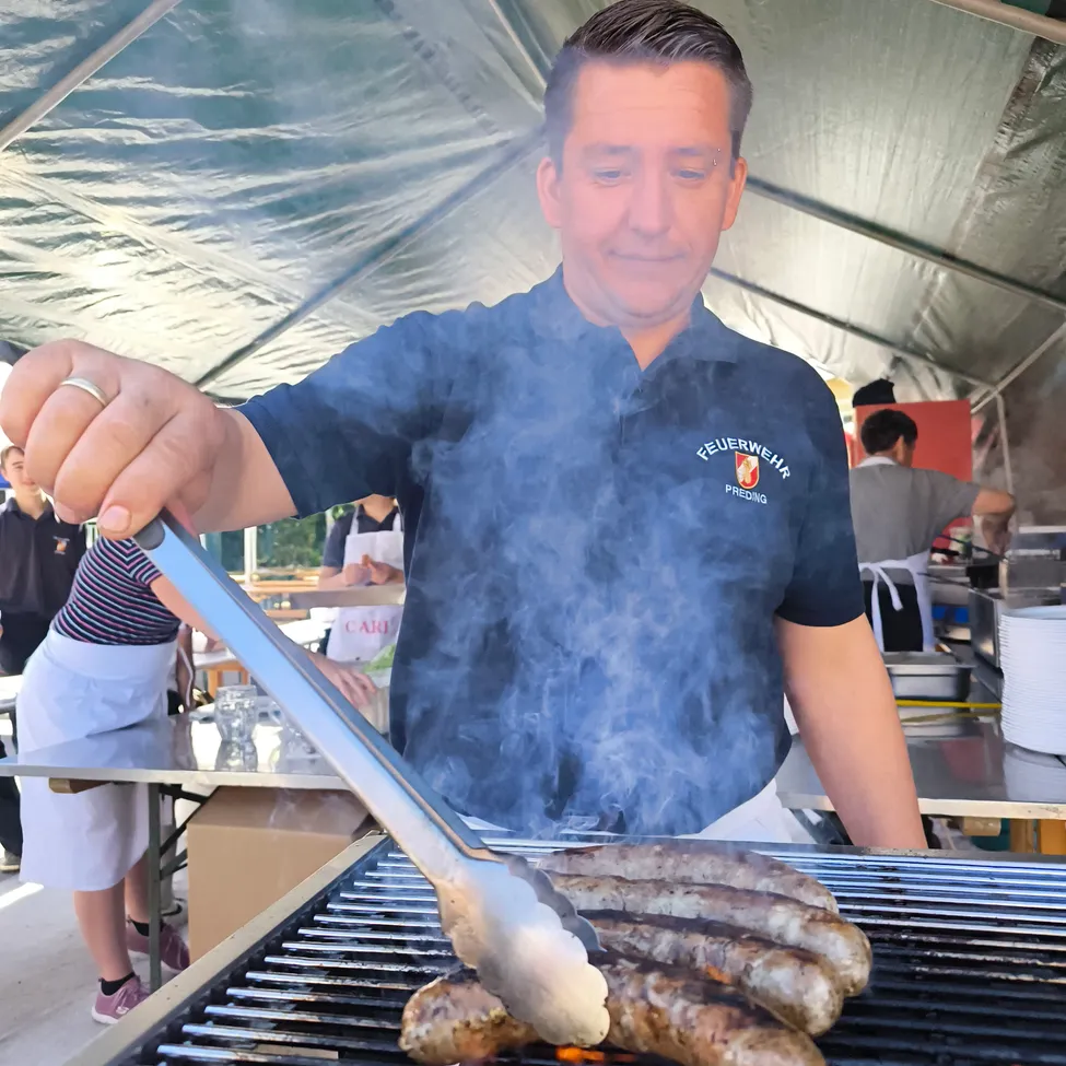 Ein Mann grillt Würste auf einem Grill unter einem Zelt. Er trägt ein blaues Shirt mit dem Wort 'Feuerwehr' darauf. Andere Menschen stehen in der Nähe.
