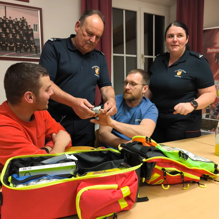 Vier Personen stehen um einen Tisch mit Erste-Hilfe-Taschen. Ein Mann in einem blauen Hemd reicht einem anderen Mann ein Werkzeug. Der Mann im roten Hemd schaut zu, während die Frau und der ältere Mann in Uniform zusehen.