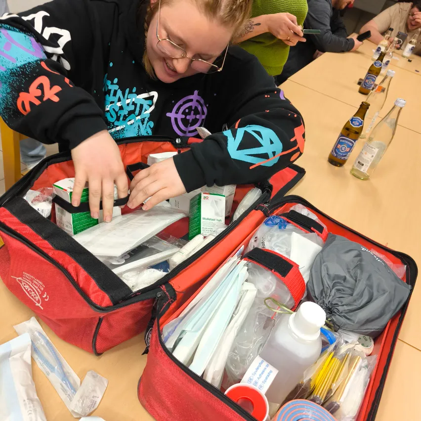 Eine Frau mit Brille öffnet eine rote Erste-Hilfe-Tasche auf einem Tisch. Verschiedene medizinische Utensilien sind darin. Hinter ihr sitzt eine Gruppe von Menschen um einen Tisch.