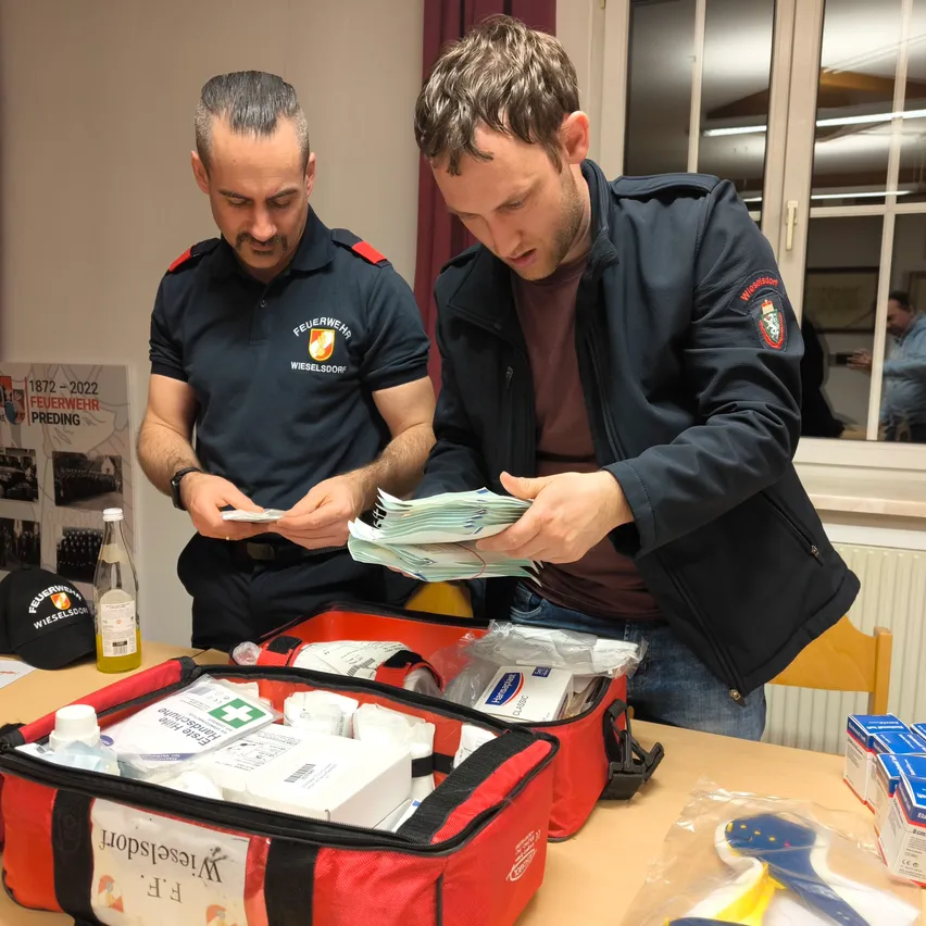 Zwei Männer untersuchen einen Bündel Geld in einem geöffneten roten Koffer auf einem Tisch. Der Mann auf der linken Seite hält ein kleines Stück Papier. Der Raum hat ein Glasfenster und ein Feuerwehr-Poster.