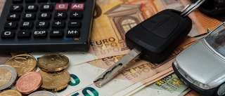 Ein Taschenrechner, Münzen und Banknoten in verschiedenen Stückelungen liegen auf einem Tisch. Ein Autoschlüssel, ein kleines Automodell und ein Stift sind ebenfalls in der Szene enthalten.