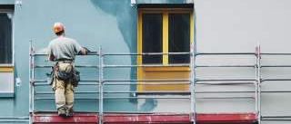 Ein Arbeiter steht auf einer Gerüst und malt ein Gebäude. Er trägt einen Helm und hat einen Pinsel in der Hand.