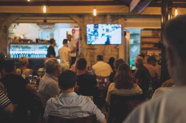 Eine Gruppe von Menschen schaut sich eine Sportveranstaltung auf einem Fernseher in einer Bar an. Die Bar hat eine Theke mit Flaschen und einen Kühlschrank.