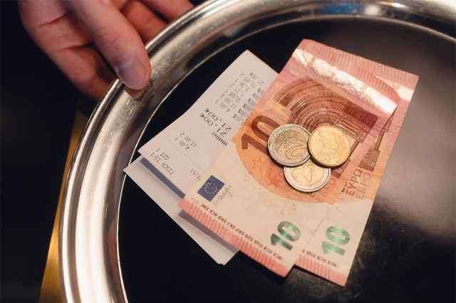 Eine Person hält ein Tablett mit ausländischer Währung und Münzen. Scheine in 10 und 20 Euro und drei Münzen sind sichtbar. Das Tablett hat eine metallische Oberfläche.