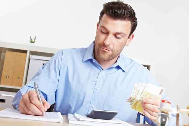 Ein Mann sitzt an einem Schreibtisch mit einem Stift und einem Notizbuch und hält Geld in der anderen Hand.