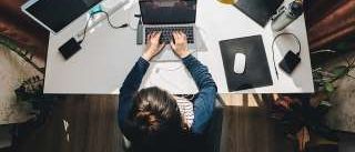 Eine Person tippt auf einem Laptop an einem Schreibtisch mit zwei Monitoren, einer Lampe, einer Mausmatte und einer Pflanze. Die Person hat Kopfhörer und ein Ladegerät in der Nähe.
