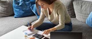 Eine Frau sitzt auf einer Couch mit Kissen und arbeitet an einem Laptop und einem Taschenrechner. Sie hat einen Notizblock, einen Stift, Geld und ein Smartphone auf dem Tisch.