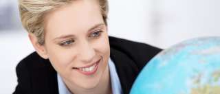 Eine lächelnde blonde Frau in einem Geschäftsanzug hält eine Erdkugel in der Hand und blickt zur Kamera.