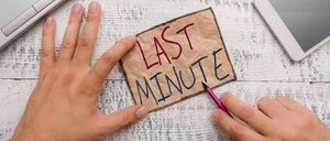 Hände halten einen Zettel mit der Aufschrift 'LETZTE MINUTE' mit einem Stift über einem Schreibtisch mit Laptop und Tablet.