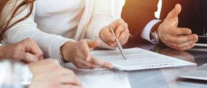 Drei Personen sitzen an einem Tisch mit einem Dokument. Eine Frau hält einen Stift und zeigt auf das Dokument, während ein Mann mit der Hand gestikuliert.