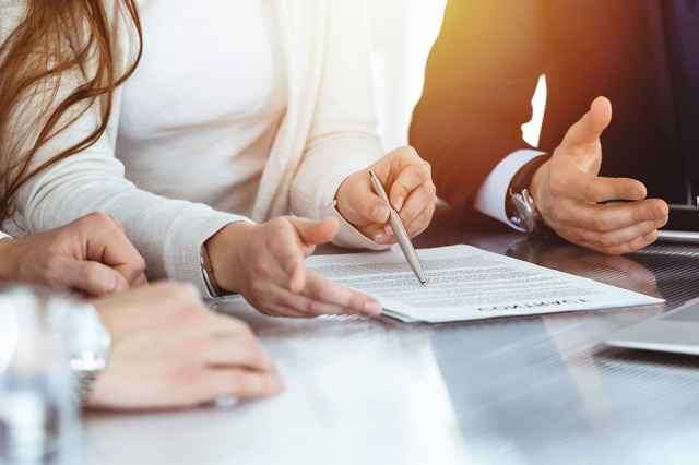Drei Personen sitzen an einem Tisch mit einem Dokument. Eine Frau hält einen Stift und zeigt auf das Dokument, während ein Mann mit der Hand gestikuliert.