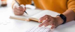 Eine Person in einem gelben Pullover sitzt an einem Schreibtisch, hält einen Stift und benutzt einen Taschenrechner, mit einem Notizbuch und einer Kaffeetasse im Hintergrund.