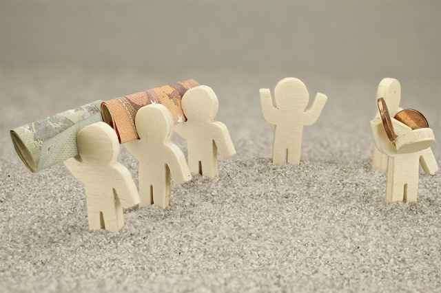 Eine Gruppe weißer Holzfiguren trägt eine aufgerollte Rechnung und Münzen auf einer grauen Oberfläche.