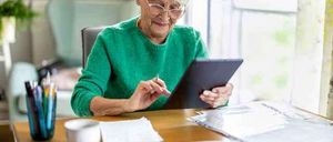An elderly woman with glasses is sitting at a table, smiling and holding a tablet. Papers, a cup, and pens are on the table.