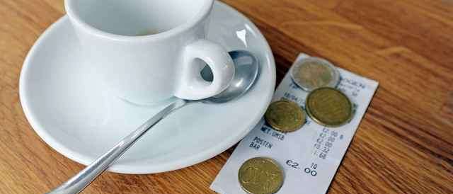 Eine weiße Tasse mit einem Löffel steht auf einer Untertasse auf einem Holztisch. Münzen und ein Bon liegen in der Nähe.