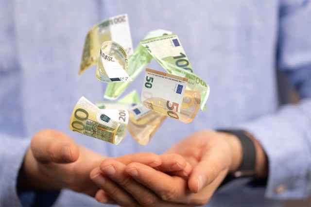 Eine Person in einem blauen Hemd fängt verstreute Euro-Banknoten mit beiden Händen auf.