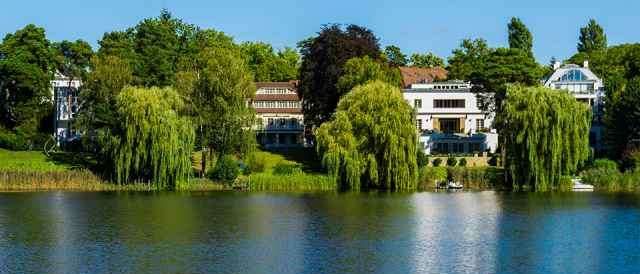 Ein ruhiger See mit Spiegelungen von Gebäuden und Bäumen. Das Ufer ist von üppiger Grünfläche gesäumt.