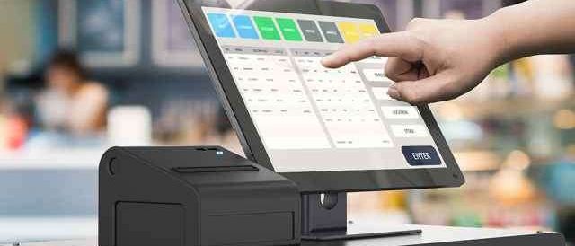 Eine Person zeigt auf einen Touchscreen-Monitor in einem Geschäft und wählt wahrscheinlich einen Artikel aus. Der Monitor zeigt ein Raster und eine Schaltfläche an.