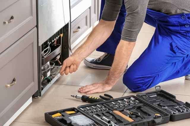 Ein Mann in blauen Hosen repariert einen Kühlschrank mit einem Werkzeugsatz auf dem Boden.