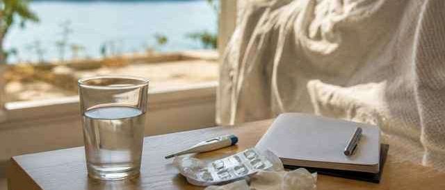 Ein Tisch mit einem Glas Wasser, einem Thermometer, Pillen, Taschentüchern, einem Notizbuch und einem Stift. Ein Bett und ein See sind im Hintergrund.