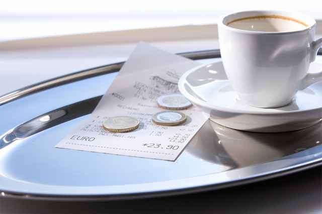 Ein silberner Tablett hält eine Tasse Kaffee, eine Untertasse, Münzen und eine Quittung. Die Quittung zeigt einen Gesamtbetrag von 23,90 Euro.