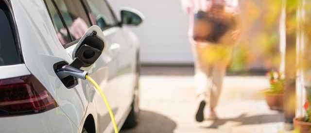 Ein weißer Elektroauto wird mit einem gelben Kabel vor einem Haus aufgeladen. Eine Frau geht im Hintergrund.