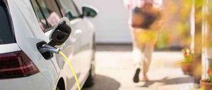 Ein weißer Elektroauto wird mit einem gelben Kabel vor einem Haus aufgeladen. Eine Frau geht im Hintergrund.
