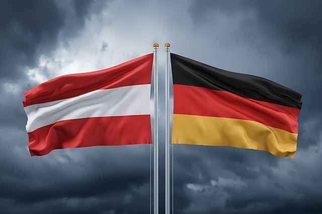 Zwei Nationalflaggen, Österreich und Deutschland, wehen im Wind gegen einen bewölkten Himmel.