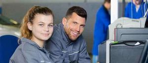 Ein Mann und eine Frau, beide in grauen Jacken, sitzen an einem Schreibtisch mit einem Computer. Der Mann lächelt und zeigt auf den Bildschirm.