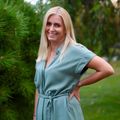 Eine blonde Frau mit einem Lächeln steht in einem Park, trägt ein türkisfarbenes Kleid und posiert mit den Händen auf den Hüften. Hinter ihr sind grüne Bäume und Gras.
