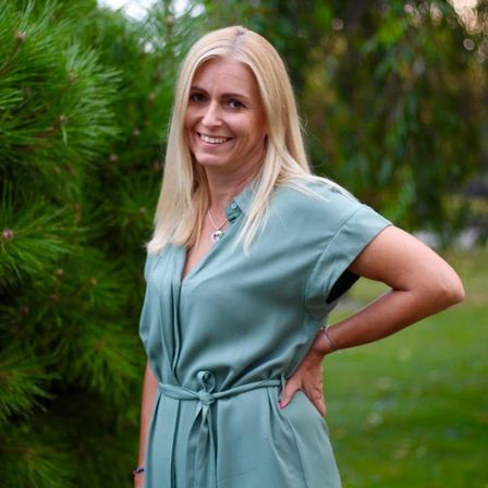 Eine blonde Frau mit einem Lächeln steht in einem Park, trägt ein türkisfarbenes Kleid und posiert mit den Händen auf den Hüften. Hinter ihr sind grüne Bäume und Gras.