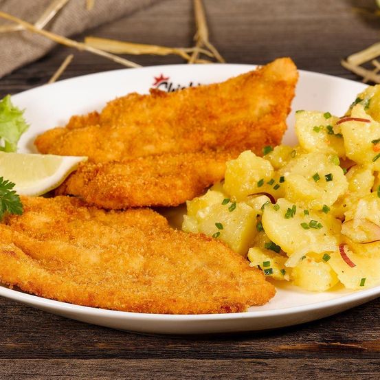 Ein weißer Teller mit gebratenem Fisch, Kartoffeln mit grünen Zwiebeln und einer Zitronenscheibe. Das Gericht steht auf einer Holzoberfläche.