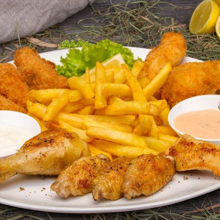 Ein weißer Teller mit gebratenen Hähnchenflügeln, Hähnchenschenkeln, Pommes und einer Schüssel Sauce. Salat, Zitrone und Brot sind auf der Seite.