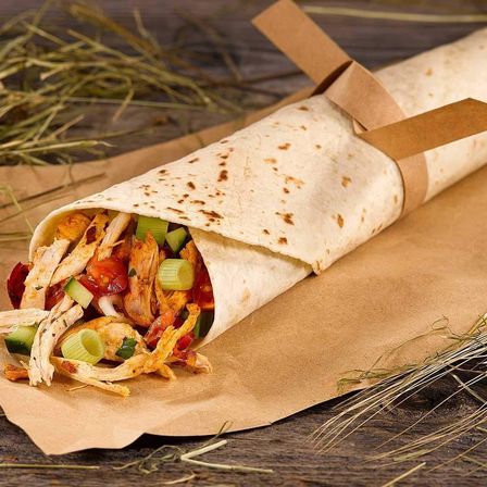 Ein Burrito mit einer weißen Tortilla gefüllt mit gehacktem Hähnchen, Tomaten und Frühlingszwiebeln, in braunes Papier gewickelt mit einer Schleife oben, ruht auf einem braunen Papierblatt auf einer Holzoberfläche.