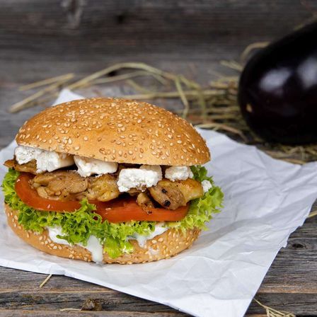 Ein Sesambrötchen-Sandwich mit Hähnchen, Feta-Käse, Tomate und Salat auf weißem Papier auf einem Holztisch mit einer Aubergine im Hintergrund.