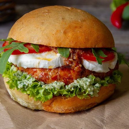 Ein Sandwich mit einem Brötchen, Mozzarella, Tomate und Basilikum liegt auf braunem Papier. Ein roter Pfeffer ist im Hintergrund sichtbar.