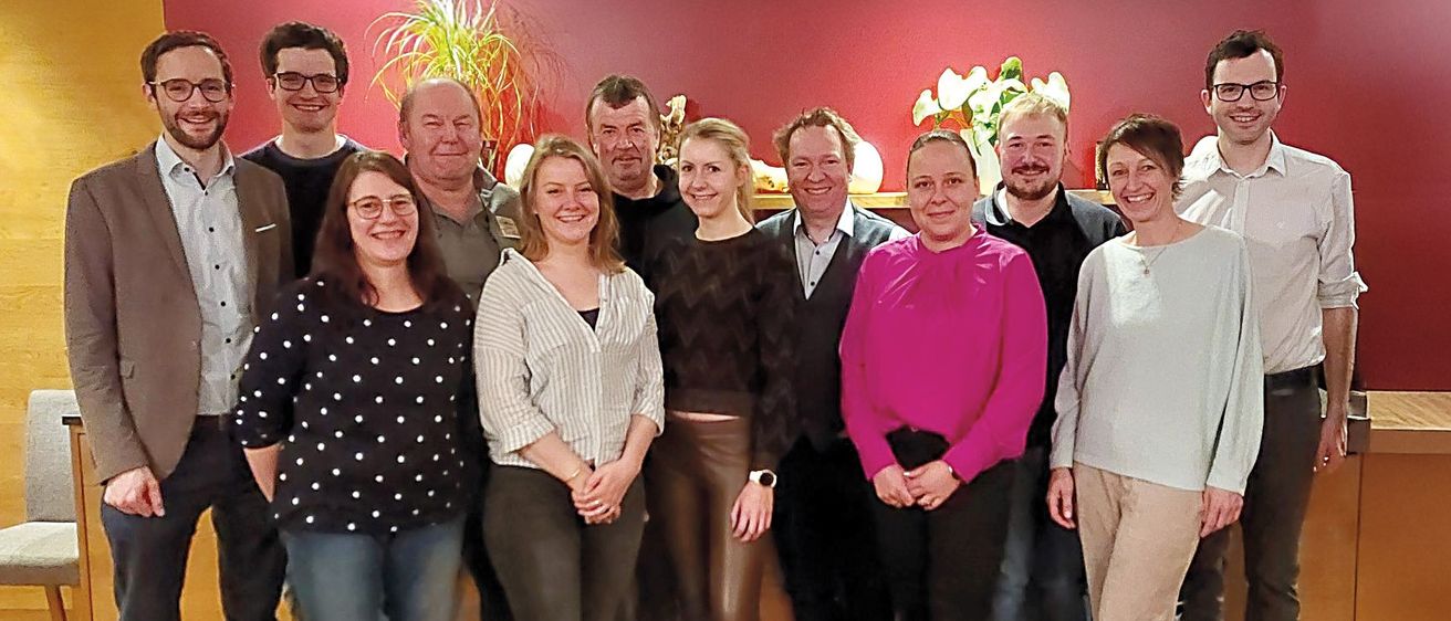 Eine Gruppe von Menschen, sowohl Männer als auch Frauen, stehen zusammen in einem Raum und lächeln für ein Foto. Sie sind lässig gekleidet, und einige tragen Brillen. Ein roter Hintergrund und ein Holzboden sind im Hintergrund zu sehen.