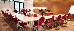 Ein leerer Konferenzraum mit einem langen Tisch und roten Stühlen. Der Raum hat Holzwände und -böden. Eine Tafel steht an der Wand.