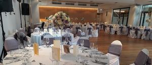 Ein Bankettsaal ist für eine Veranstaltung dekoriert mit runden Tischen, weißen Tischdecken und Blumen-Zentren. Stühle sind um die Tische herum angeordnet, und einige haben dekorative Schärpen.