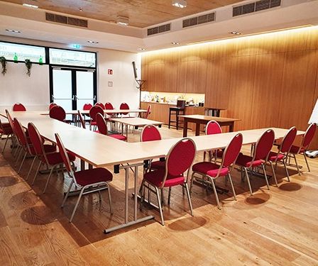 Ein leerer Konferenzraum mit langen Tischen und roten Stühlen. Holzwände und -böden schaffen eine warme Atmosphäre. Eine Whiteboard steht rechts.