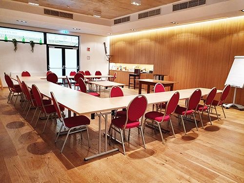 Ein leerer Konferenzraum mit langen Tischen und roten Stühlen. Holzwände und -böden schaffen eine warme Atmosphäre. Eine Whiteboard steht rechts.