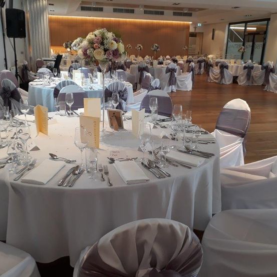 Ein wunderschön dekorierter Bankettsaal mit für eine Veranstaltung gedeckten Tischen. Jeder Tisch hat einen Blumenschmuck, weiße Tischdecken und ordentlich arrangierte Bestecke. Die Stühle sind mit Seidenschleifen geschmückt.